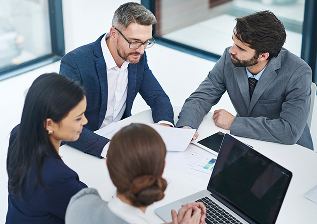 Vier Personen, die mit einem Laptop an einem Meeting-Tisch sitzen und etwas besprechen